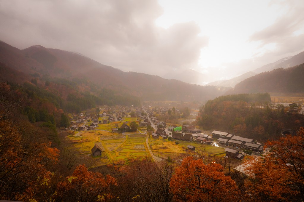 A scenic view of a mountainous village surrounded by colorful autumn foliage, under a cloudy sky with hints of sunlight illuminating the landscape.