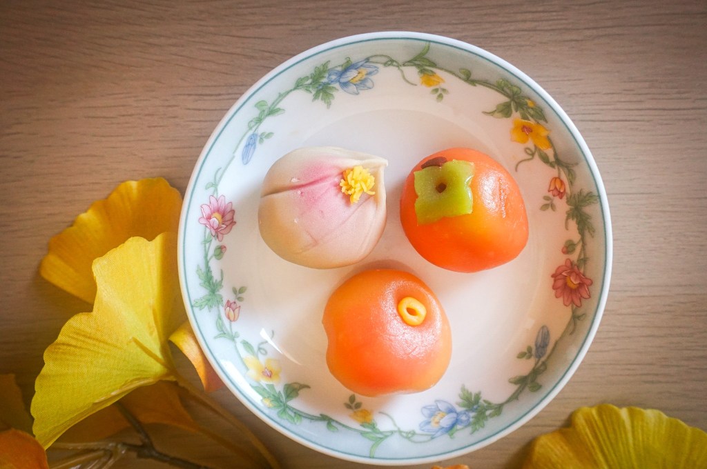 A decorative plate with three colorful, elaborately shaped Asian sweets resembling fruits, surrounded by yellow leaves.